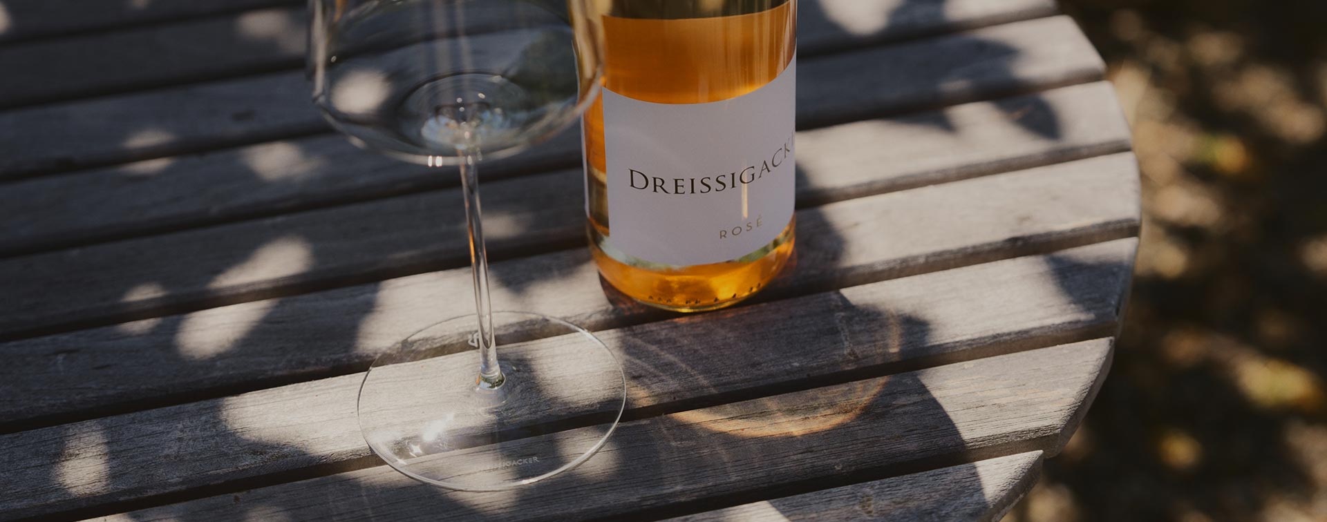 Flasche Dreissigacker Rosé mit leerem Glas auf rustikalem Holztisch im Schatten eines Baumes